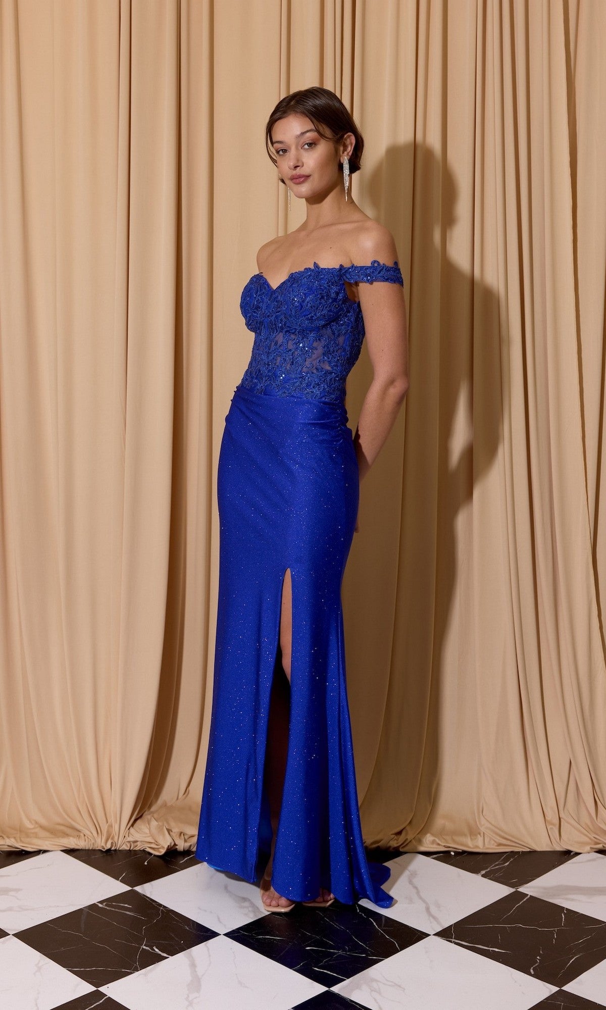 A woman stands on a black and white checkered floor in front of beige curtains, wearing the Eureka EK173 royal blue off-the-shoulder long prom dress with a lace bodice and thigh-high slit.